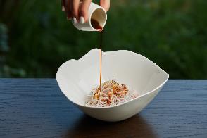 A person pouring sauce into a bowl of noodles

Description automatically generated